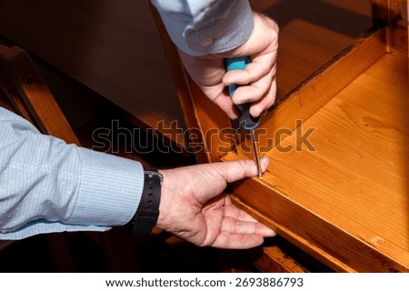 A man is doing carpentry work in his house. He holds the chair with one hand to keep it from moving. With the other hand he holds a hand screwdriver 