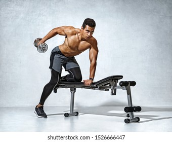 Man doing exercise with dumbbell leaning on sports bench. Photo of muscular man with naked torso and good physique on grey background. Strength and motivation