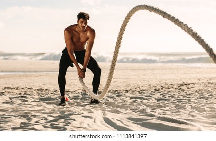 Man Doing Fitness Workout At A Beach On A Sunny Day. Bare Chested Man Doing Workout Using A Battle Rope On The Beach.