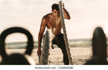 Man doing fitness workout at a beach on a sunny day. Bare chested man doing workout using two battle ropes on the beach with kettlebells in the foreground.
