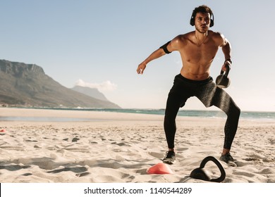 Man doing fitness workout at a beach using a kettlebell. Bare chested athletic man doing exercise with kettlebells wearing wireless headphones and mobile phone fixed to armband.