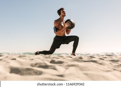 Man doing fitness workout at a beach using a medicine ball. Bare chested athletic man doing stretches holding a medicine ball wearing wireless headphones and mobile phone fixed to armband. - Powered by Shutterstock