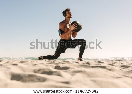 Man doing fitness workout at a beach using a medicine ball. Bare chested athletic man doing stretches holding a medicine ball wearing wireless headphones and mobile phone fixed to armband.
