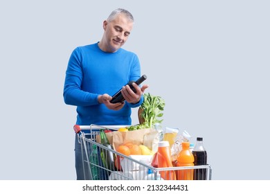 Man doing grocery shopping at the supermarket, he is buying a bottle of red wine, full shopping cart in the foreground - Powered by Shutterstock