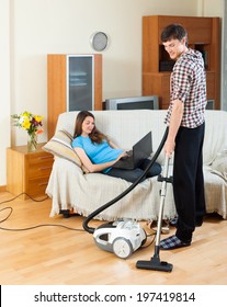 Man Doing House Cleaning With Vaccuum Cleaner While Girl  With Laptop  Resting Over Sofa