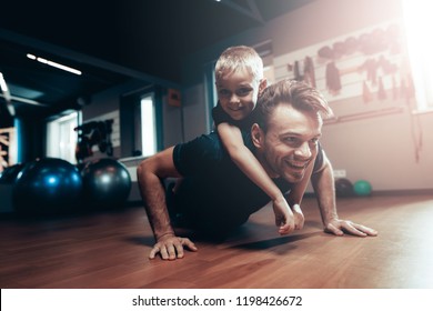 Man Is Doing Push Ups In Gym With Son On A Back. Parenthood Relationship. Sporty Family Concept. Active Lifestyle. Triceps Exercise. Holiday Leisure. Working Out Together. Fitness Day.