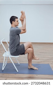 Man doing seated variation of eagle pose sitting on metal chair indoor. Yoga Asana with focus on arms relieves tension in neck and shoulders and stretch the upper back