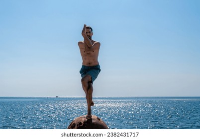 Man doing yoga exercise outdoors standing on old rusty floating marine mine on the beach with rocky shore and sea background. Healthy lifestyle, pollution, nature protection, war and peace concept. - Powered by Shutterstock