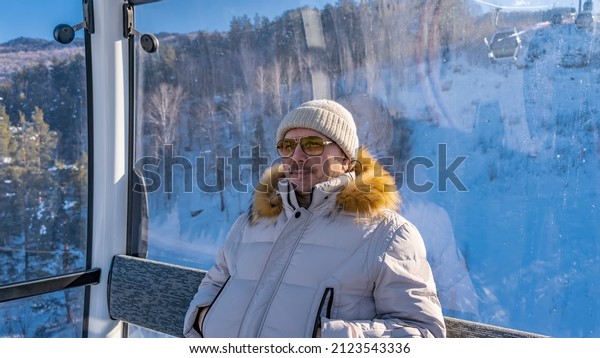 A man in a down jacket, hat, sunglasses is\
sitting in a funicular cab. He looks into the distance and smiles.\
A snow-covered wooded mountain slope is visible behind the glass.\
Altai. Manzherok