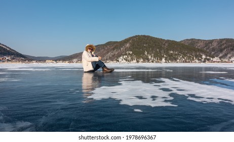 A man in a down jacket and a hood sits on a frozen lake. The legs are bent at the knees. Reflection on ice. Around - areas of snow. On the shore there are wooded mountains against the blue sky. Baikal