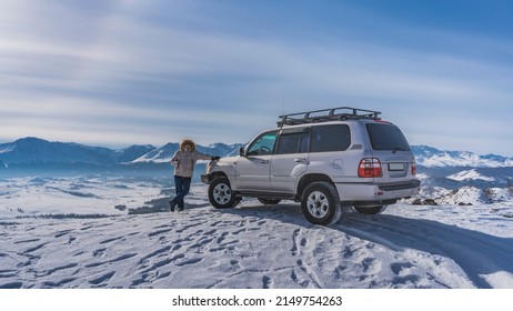 A man in a down jacket stands on the edge of a snowy plateau, leaning on an SUV. Footprints in the snow. In the distance - a mountain range, a valley. Blue sky. Altai - Shutterstock ID 2149754263