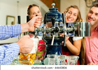 Man Drawing A Beer From Tap On A Kegerator In Pub Or Inn, People In Bavarian Tracht Are Standing In The Background