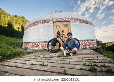 Man with dreadlocks and beard sitting in front of mountain bike with tourist bags near Nomad Yurt house in beautiful mountain valley in Almaty, Kazakhstan. Extreme Sport bicycle bikepacking  travel - Powered by Shutterstock