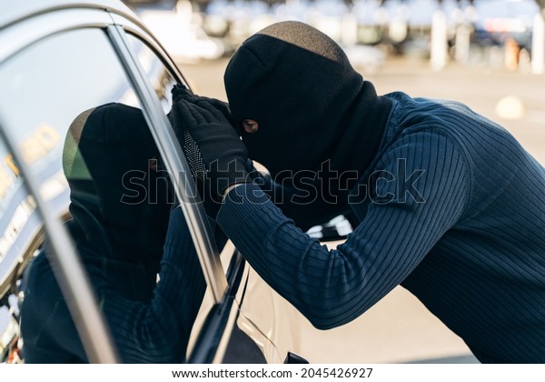 Man dressed in black with a balaclava on his head\
looking at the glass of car before the stealing. Car thief, car\
theft concept 