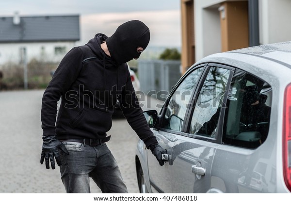Man dressed in black\
with a balaclava on his head pulls the handle of a car. Car thief,\
car theft concept
