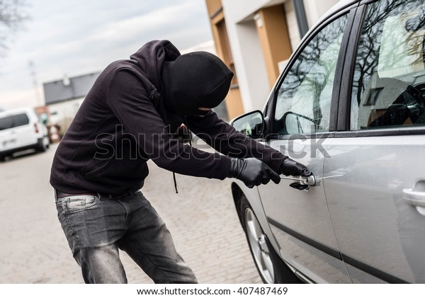 The man dressed in black with a balaclava on his\
head trying to break into the car. He uses a screwdriver. Car\
thief, car theft concept