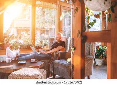 Man Dressed Black T-shirt And Jeans Sitting In A Comfortable Armchair, Using A Modern Slim Laptop And Drinking Tea In House Sunroom Living Room. Distance Or Freelance Or Writer Working Concept Image.