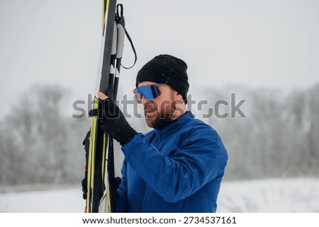 Man dressed in blue winter jacket and black beanie holds ski poles in a snowy environment with trees visible in the background