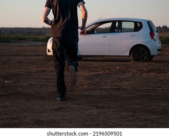 Man Dressed In Dark Clothes Running Towards A White Car On A Dirt Road With The Early Morning Light