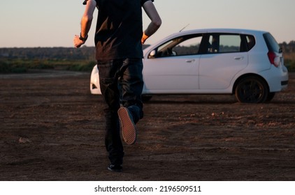 Man Dressed In Dark Clothes Running Towards A White Car On A Dirt Road With The Early Morning Light