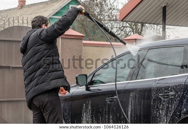 A man\
dressed in dark clothes washes his car in his yard, using a high\
pressure washer. The car is covered with\
foam.