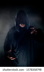 Man Dressed In A Dark Robe Looking Like A Cult Leader On A Smoky Or Foggy Background.  He Looks Like A Creepy Evil Villain