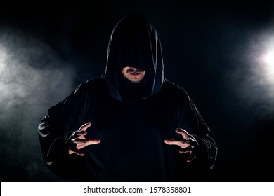 Man dressed in a dark robe looking like a cult leader on a smoky or foggy background.  He looks like a creepy evil villain