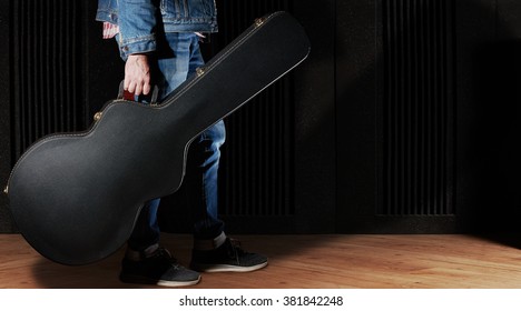 Man Dressed In Jeans Holding Guitar Case Against Dark Background