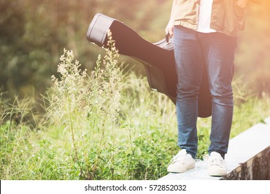 Man Dressed In Jeans Holding Guitar Case In Nature Outdoor.