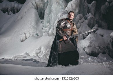 Man Dressed In Medieval Armor And Raincoats With Swords In Winter In The Mountains Under Heavy Snow Blizzard. Fantasy Warrior Knights In Winter Snowy Landscape