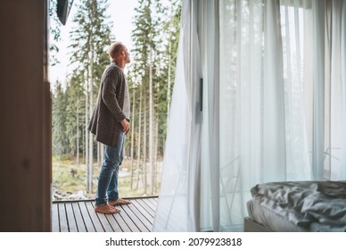 A Man Dressed Open Knitted Cardigan Standing On A Forest House Balcony And Enjoying The Fresh Air With Nature Forest Pine Trees View. Outside Scandinavian Interior Design House. Living In Wild Concept