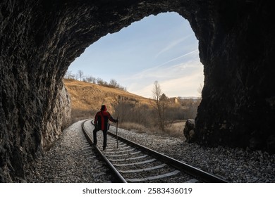 a man dressed in a red jacket stands at the end of an old railway tunnel and looks at the beautiful mountain landscape in front of him, holding a large wooden stick in his hand.
