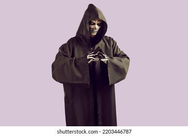 Man dressed in scary Halloween costume. Studio portrait of Mr Death with skeleton skull makeup on face wearing black cape with hood looking at camera while standing isolated on light purple background - Shutterstock ID 2203446787