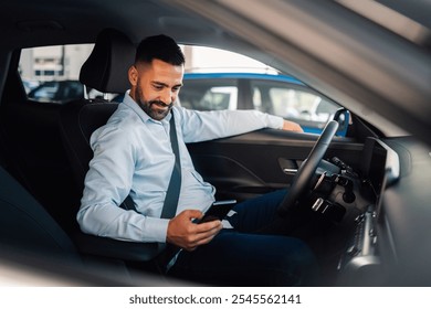 A man, dressed smartly, is sitting inside a car looking at his smartphone and connecting it with his new car with a smile, suggesting satisfaction and connectivity in a modern urban setting. - Powered by Shutterstock