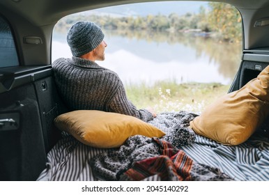 Ein Mann mit warmen Strickbekleidung sitzt im gemütlichen Camper-Kofferraum und genießt den Blick auf den Bergsee. Warmes Frühherbst Auto-Reisen Konzept in Pkw-Piloten und Decken-Blick Bild.  