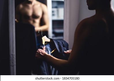 Man Dressing Up. Guy Choosing Suit And Tie, Elegant Clothes And Outfit. Businessman Standing In Front Of A Mirror And Looking At His Reflection. Young Fit Shirtless Person Getting Ready In Dark Room.