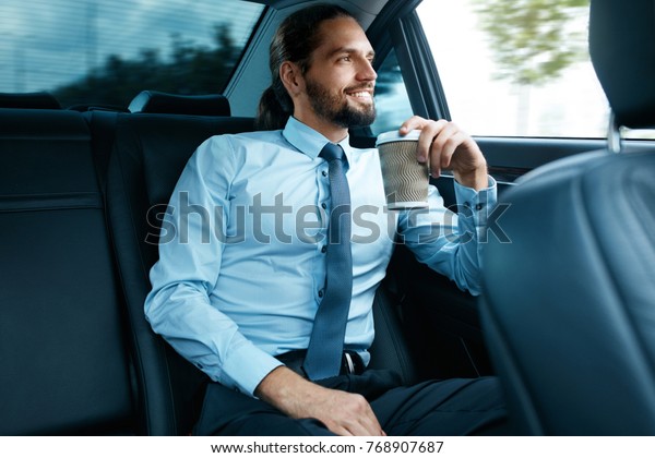 Man Drinking Coffee In Car. Portrait Of\
Successful Confident Smiling Businessman Drinking Hot Drink In Cup,\
Looking Through Window While Riding On Back Seat Of Car, Going To\
Work. High Resolution
