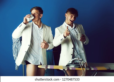 Man drinking from cup in dressing room. Macho winking and pointing finger. Businessmen wearing shirts, jackets and underpants. Fashion. Ironing board with iron and clothes rack on blue background.
