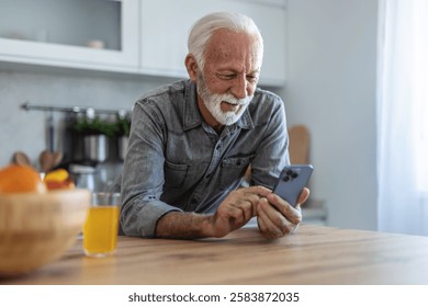 Man drinking orange juice and using smart phone. Elderly man, pensioner, sitting in kitchen and using smartphone, he is in nursing home or in his apartment, concept of aging and happy retirement days.