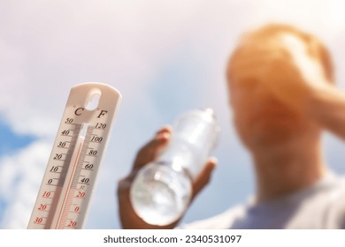 Man drinking water in extreme heat, Thermometer in summer day shows or indicate high temperature degree with sun in background. - Shutterstock ID 2340531097