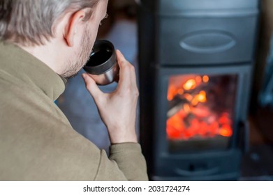 A man drinks hot tea from a thermos in front of a burning fireplace at home. Close-up. Country house, winter, rest, relaxation. Subject is out of focus  - Powered by Shutterstock