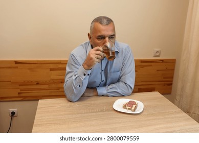 Man drinks tea at a kitchen table beside a sandwich, spending a quiet moment alone at home.