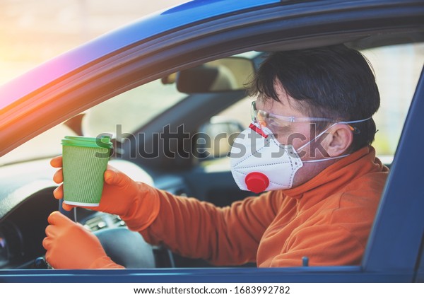 A\
man driver in medical face mask and safety glasses during an\
epidemic in the car. The man drinks a coffee in the\
car
