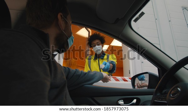 Man driver in safety mask stopping near security\
booth for temperature scanning before entering area. Afro security\
worker in uniform and protective mask measuring driver temperature\
before open gates