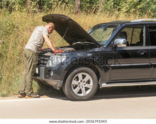 Man driver\
standing and looking inside hood raised up and thinking about\
problem after car break in\
trip.