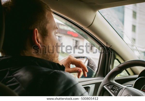 man drives a car.\
A guy in a jacket sits behind the wheel of a car traveling on road\
in traffic jam, smoking a cigarette. Photo taken off the seat, due\
to the driver\'s back