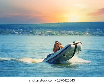 Man Drives An Inflatable Motor Boat In A Lake Or Sea At Sunset. The Boat Makes A Sharp Turn And Splashes And Waves Go In All Directions