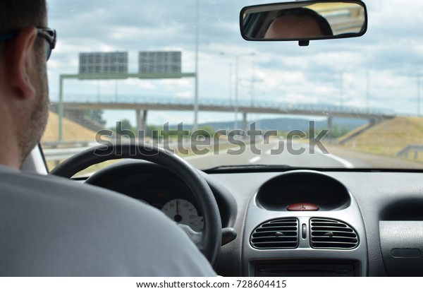 Man\
driving a car with a dirty front glass after a long drive on a\
highway with roadsigns and a viaduct  in front of\
him