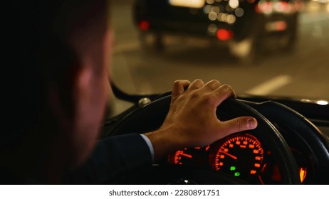 Man driving car fast in night city street with traffic. Road trip, speeding, driver concept. Commuter, male taxi cab driver. Interior back view of male hand on steering wheel in car. Close up