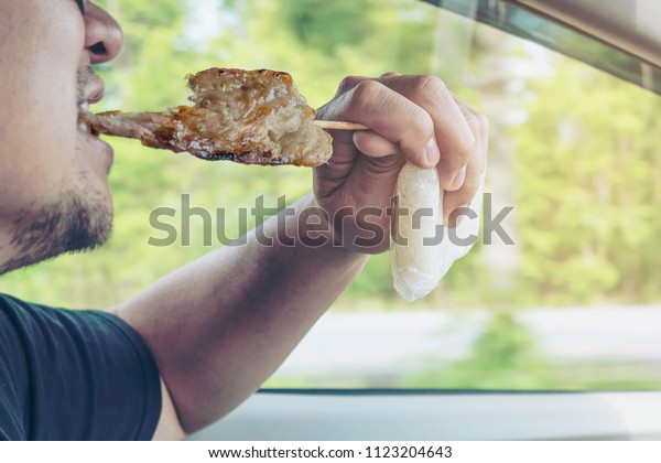Man driving car while eating grilled  pork stick\
with sharp spike bamboo stick and sticky rice in plastic pack\
dangerously - multitasking driving bad behave danger risk drive\
concept